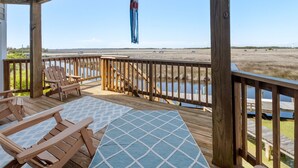 Eine Holzterrasse mit Liegestühlen und einem Tisch, mit Blick auf einen Strand und ein Gewässer.