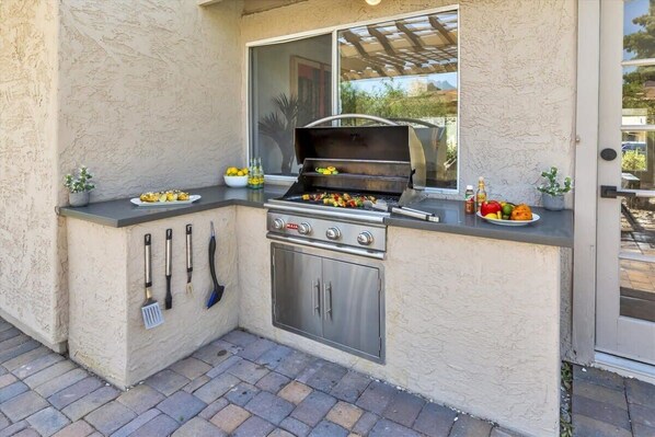 An outdoor kitchen with a grill, cooking utensils, and a window overlooking a garden. - Spacious Desert Home, Heated Pool (Scottsdale)