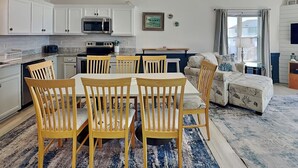 Una cucina moderna con mobili bianchi, un forno a microonde e un piano cottura. È presente una zona pranzo con tavolo e sedie. In zona soggiorno si vedono un divano e una finestra con tende.