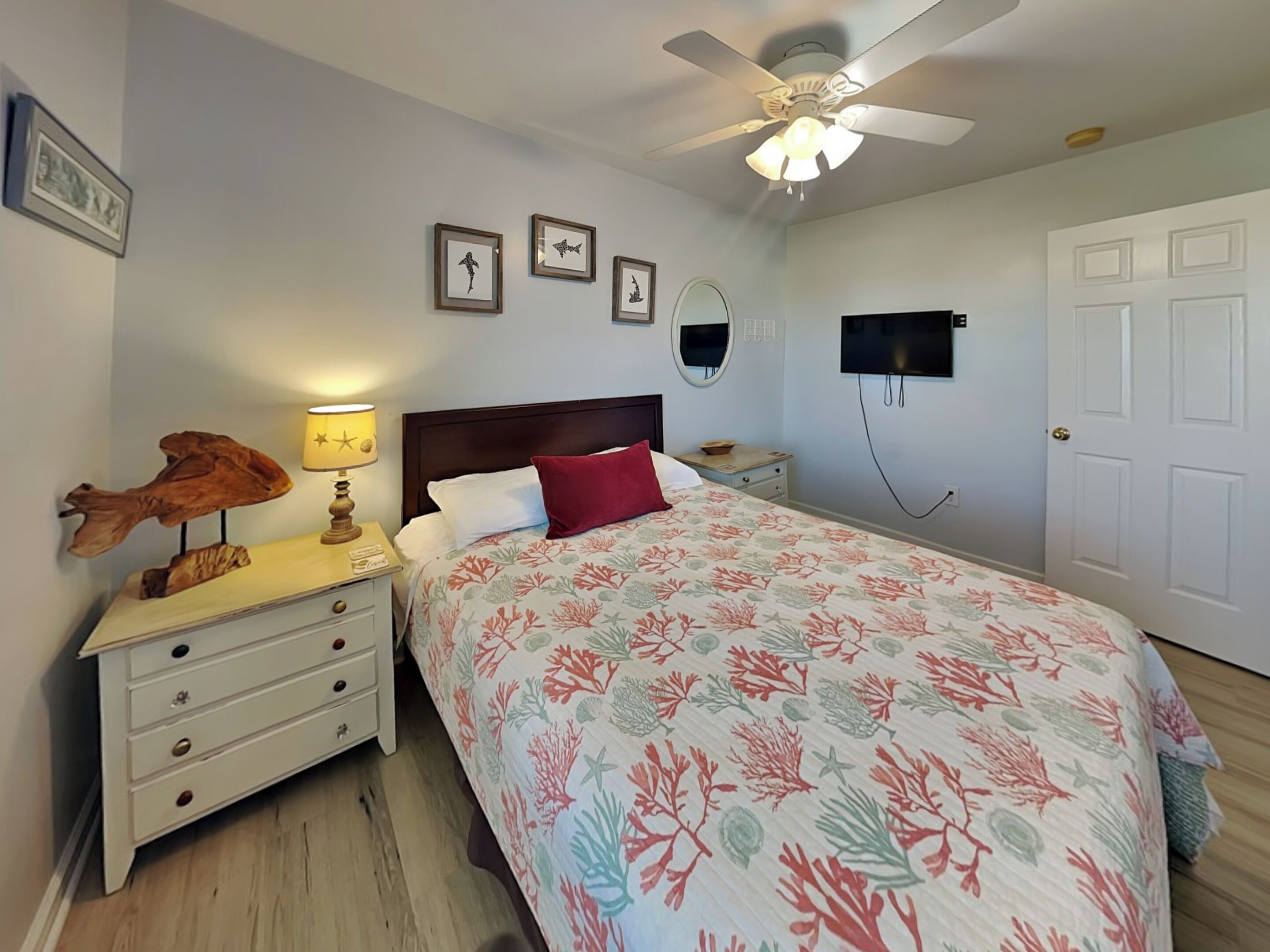 A bedroom with a bed, nightstand, lamp, framed pictures, ceiling fan, TV, and a door.