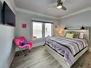 Un dormitorio con cama, ventilador de techo, ventana, silla y televisor fijado a la pared.