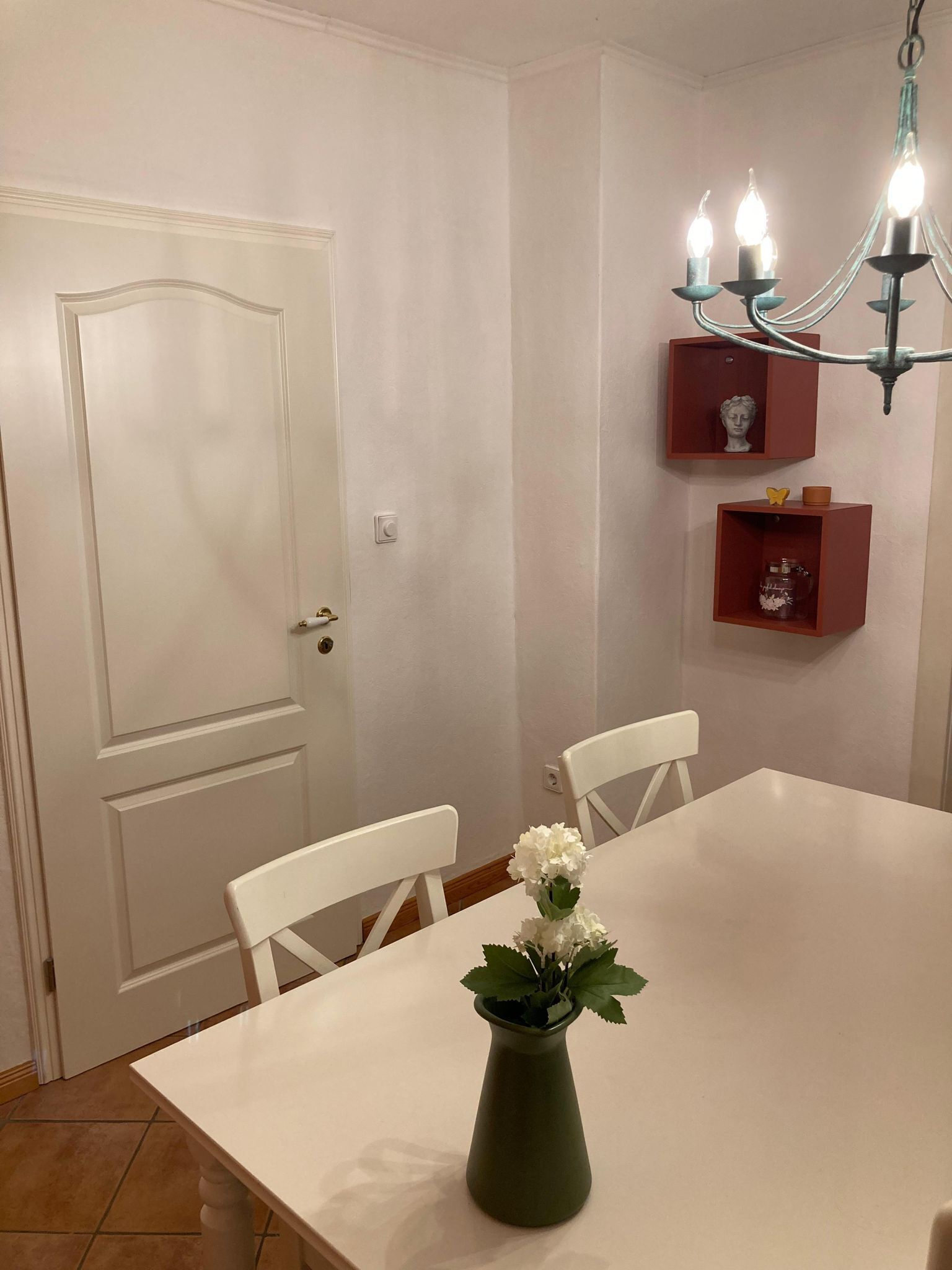 Ein Essbereich mit einem weißen Tisch, zwei weißen Stühlen, einer grünen Vase mit Blumen und einer Kristallleuchter.