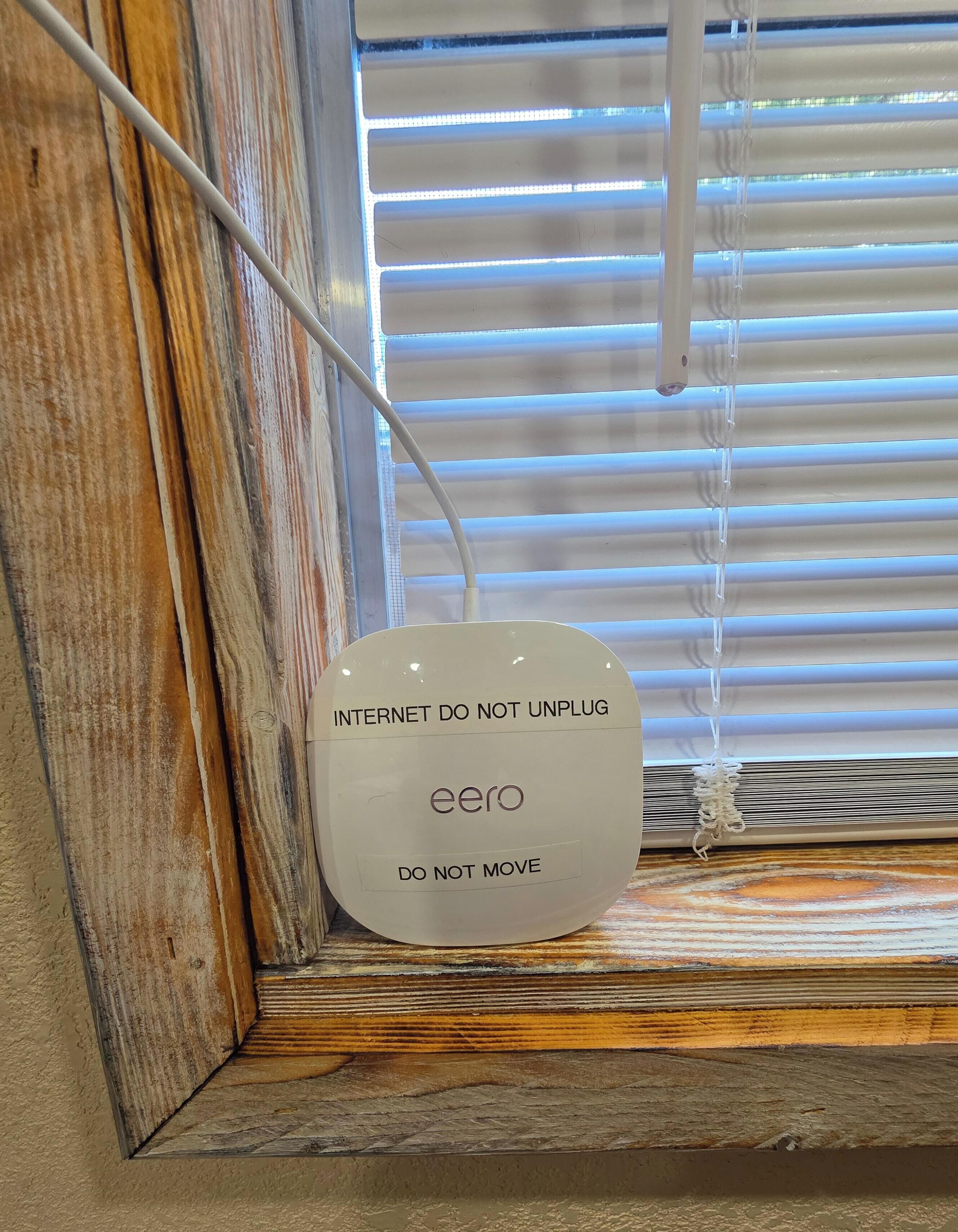 A device labeled 'eero' with 'INTERNET DO NOT UNPLUG' and 'DO NOT MOVE' instructions, placed on a wooden ledge near a window with blinds.
