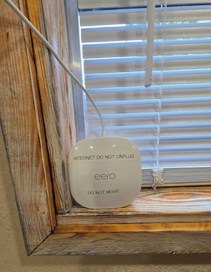A device labeled 'eero' with 'INTERNET DO NOT UNPLUG' and 'DO NOT MOVE' instructions, placed on a wooden ledge near a window with blinds.