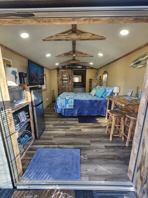A compact RV interior with a kitchenette, a bed with a blue bedspread, a dining area with stools, and a ceiling fan.