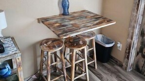A rustic dining area with a wooden table and two stools, a decorative fish wall art piece, and a surfboard sign. - The Sea Shed (Perry)