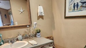 A bathroom with a white sink, vanity, and toilet. There is a starfish decoration on the wall, a framed picture above the toilet, and a small plant on the vanity. - The Sea Shed (Perry)