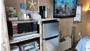 A compact kitchenette with a microwave, toaster, and refrigerator. A television is mounted on the wall above the counter, and a starfish decoration hangs on the wall. - The Sea Shed (Perry)