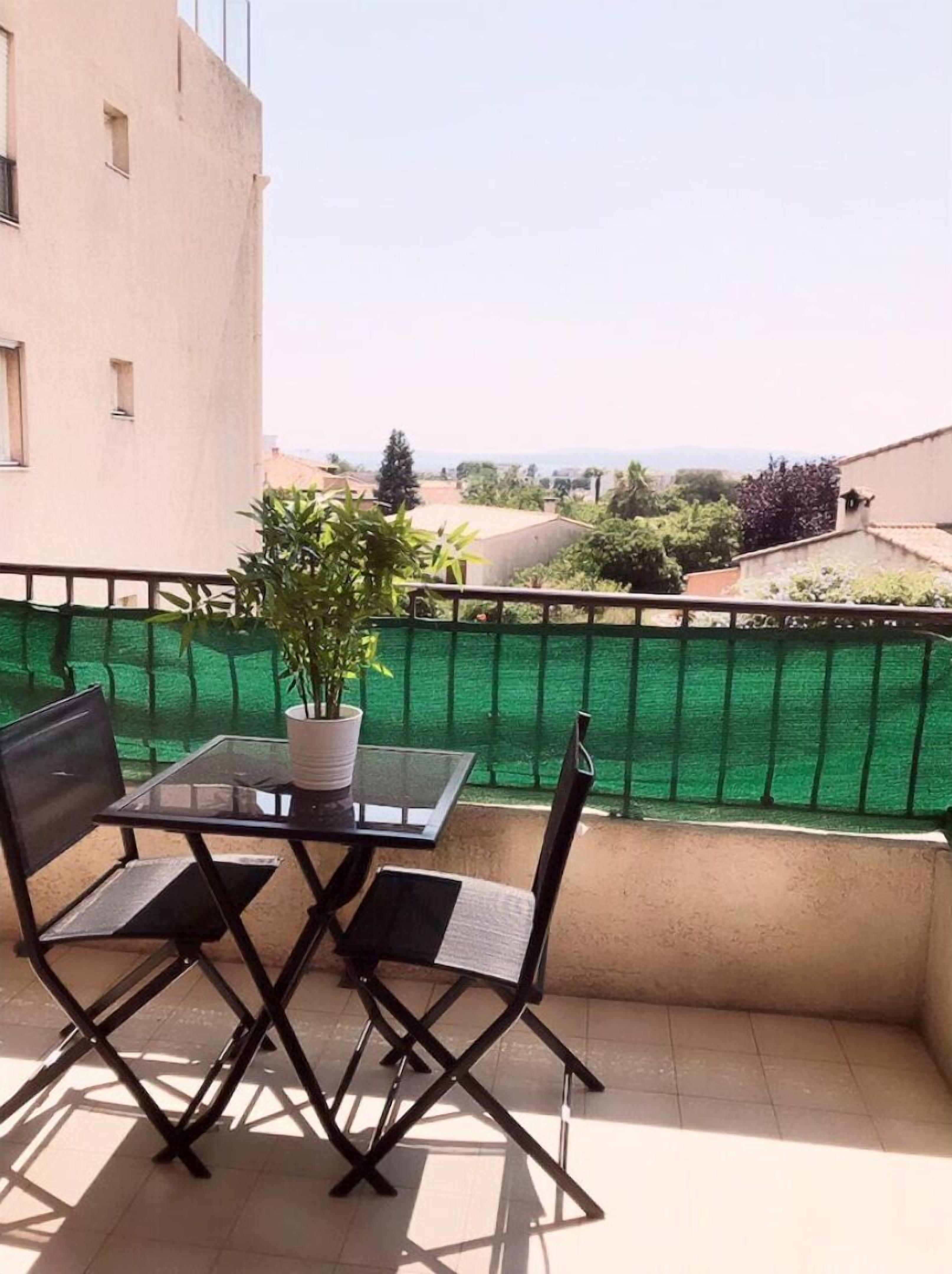 Un balcone con un tavolino in vetro e due sedie, una pianta in vaso e un recinto verde.