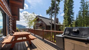 Un terrazzo in legno con tavolo da picnic e barbecue, con vista su una baita e una foresta.