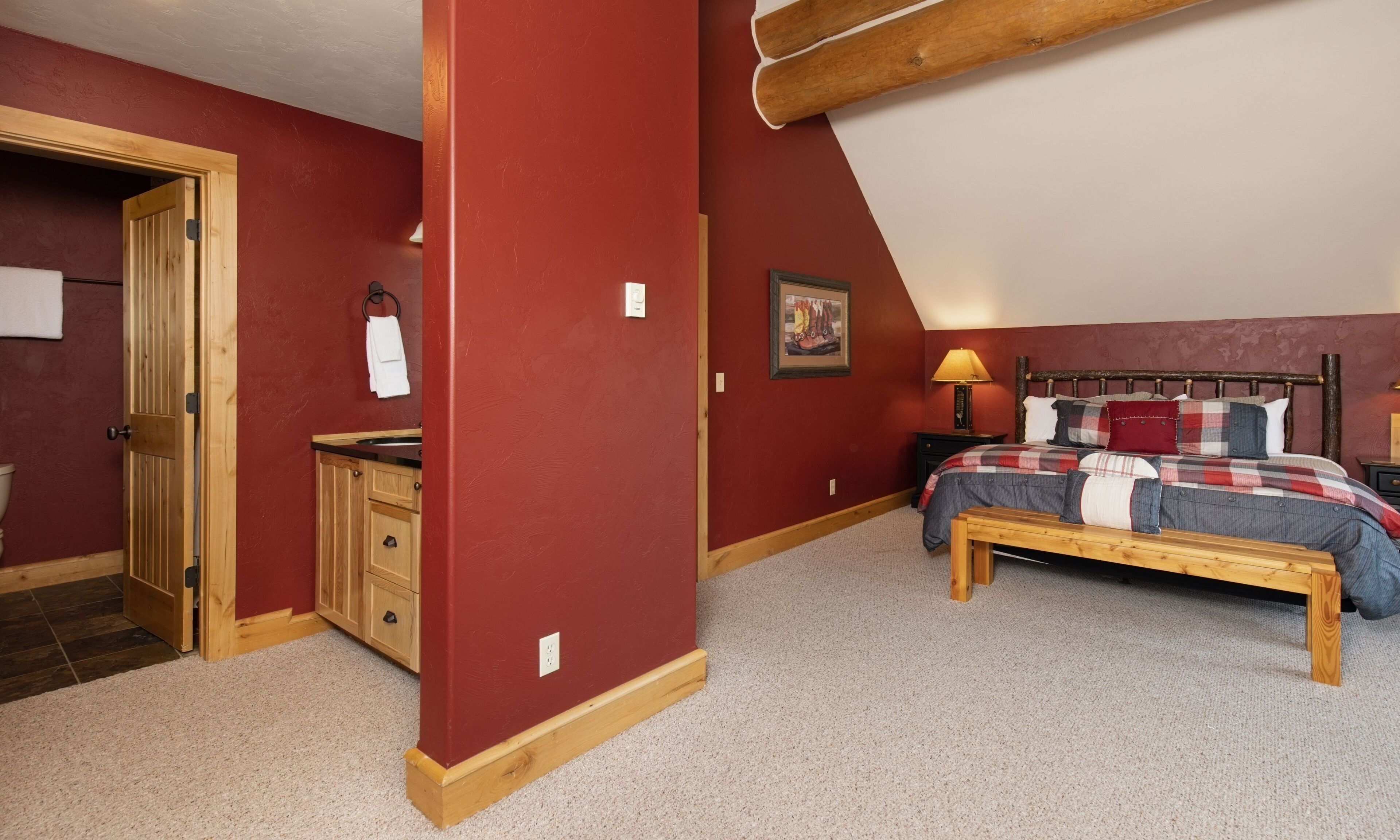 A bedroom with a wooden bed, a bench, a nightstand, a painting, and a door leading to another room.