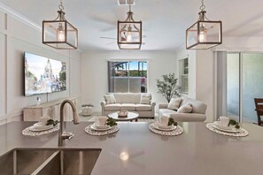 A modern kitchen with a central island, a dining area with white furniture, and a large window providing a view of the outdoors.