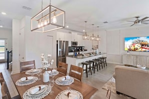 A modern kitchen with stainless steel appliances, a dining area with a wooden table set for four, and a living area with a sofa and TV.