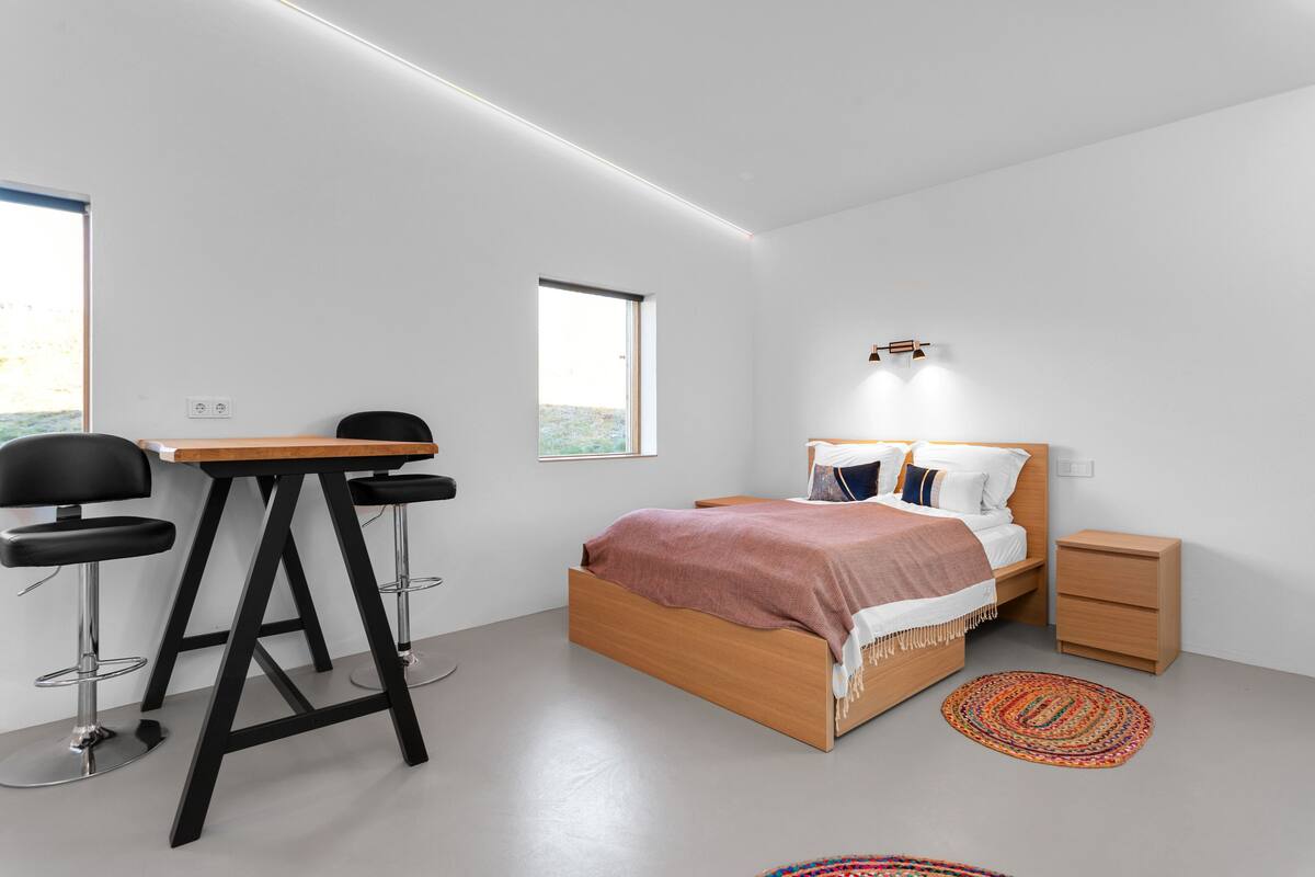 A modern bedroom with a wooden bed, a small bedside table, a chair, and a desk with stools.