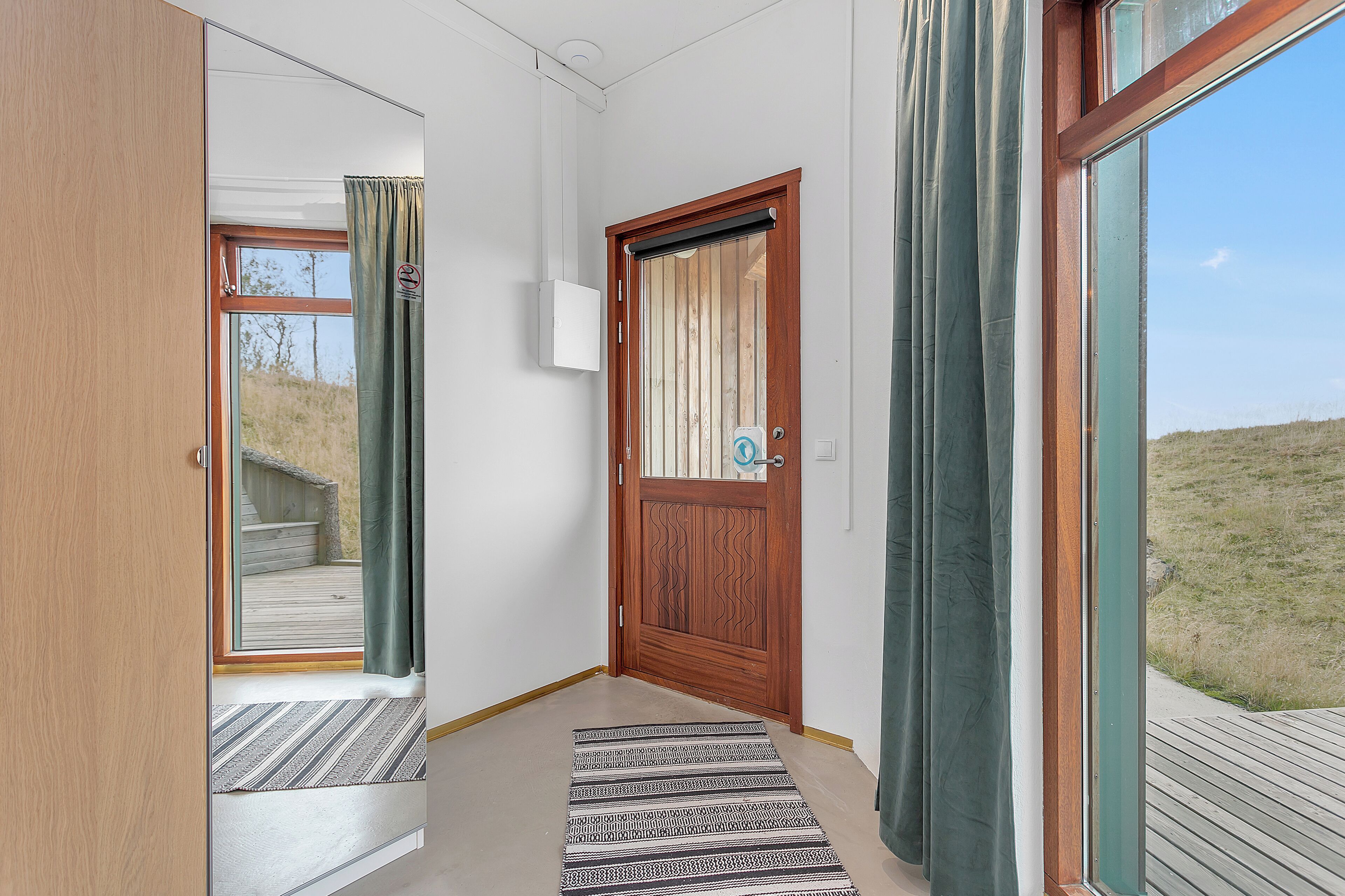 A hallway with a wooden door, a window with curtains, and a view of a grassy area outside.