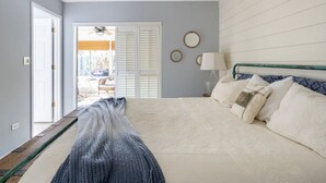 A bedroom with a large bed, a ceiling fan, a window with blinds, and a door leading to an outdoor area.