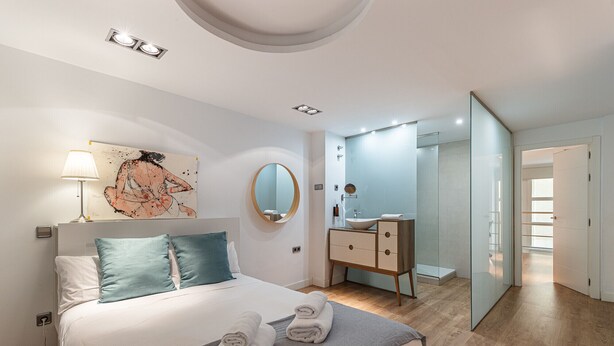 A modern bedroom with a bed, a bedside lamp, a painting, a round mirror, a vanity with a sink, and a glass door leading to another room. - Waou Chamberi VII - Alquiler Corta Duración (Madrid)