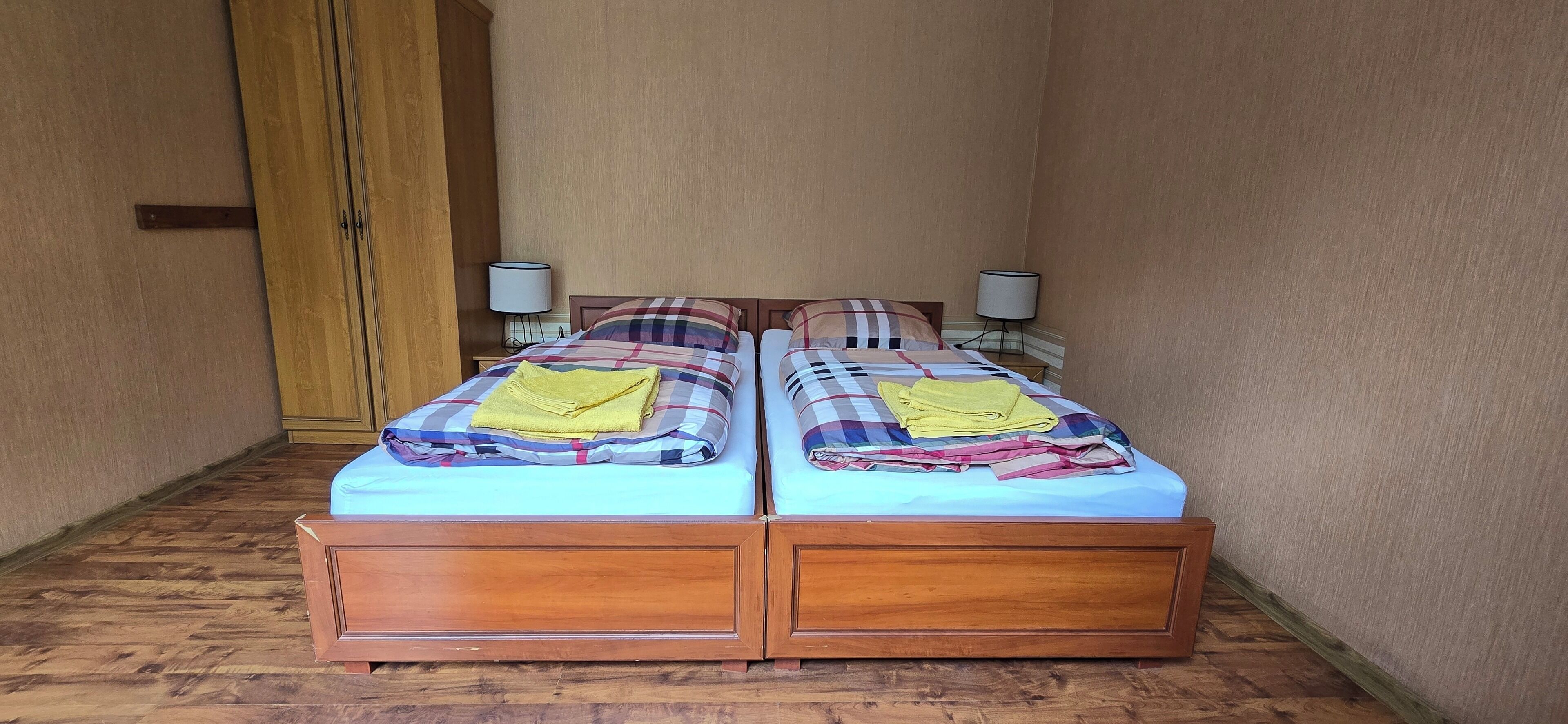 A room with two wooden beds, each with a plaid bedspread and a yellow towel. There are two bedside tables with lamps.