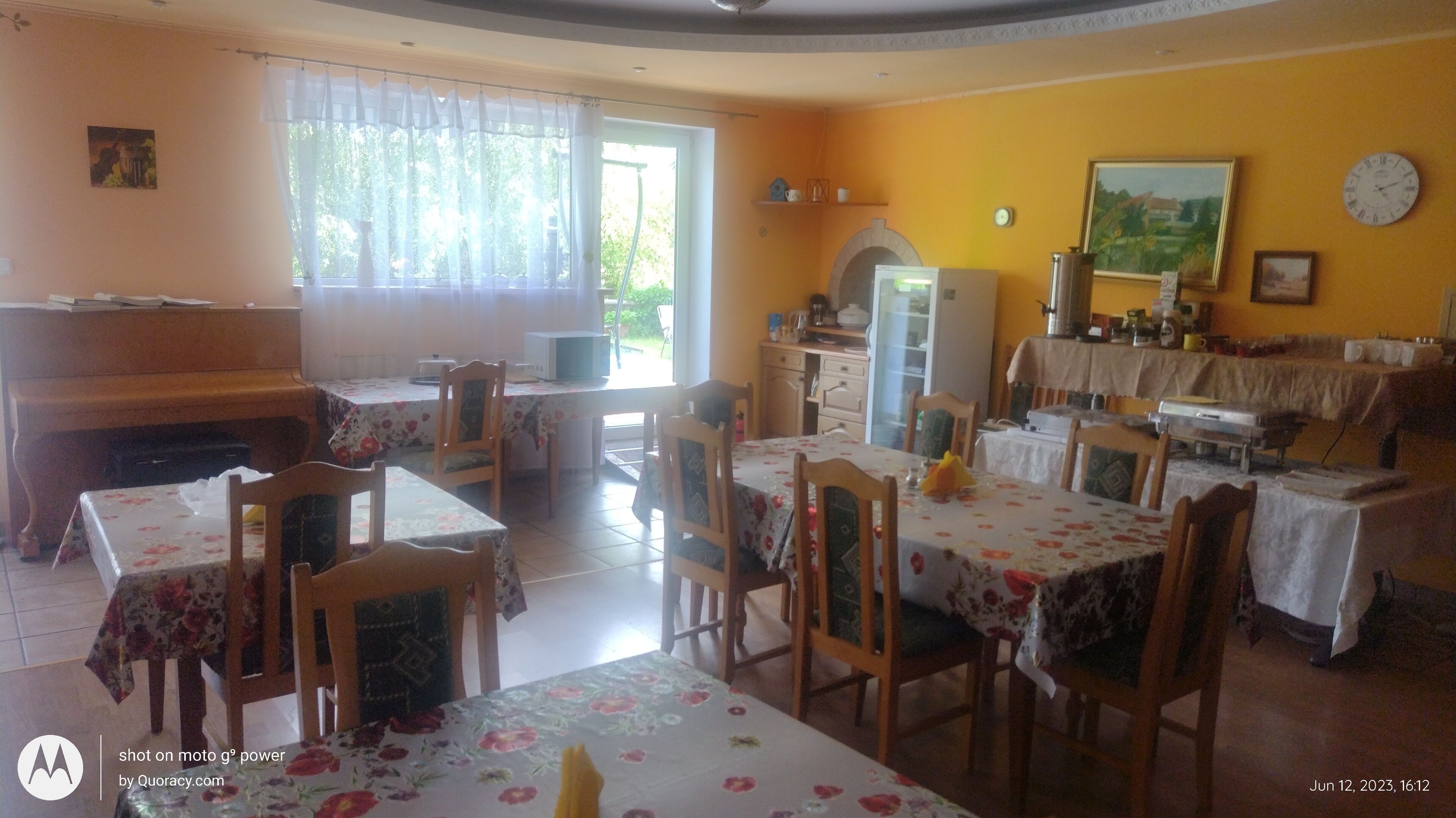 A dining area with multiple tables and chairs, a piano, and a kitchen area with a refrigerator and microwave.