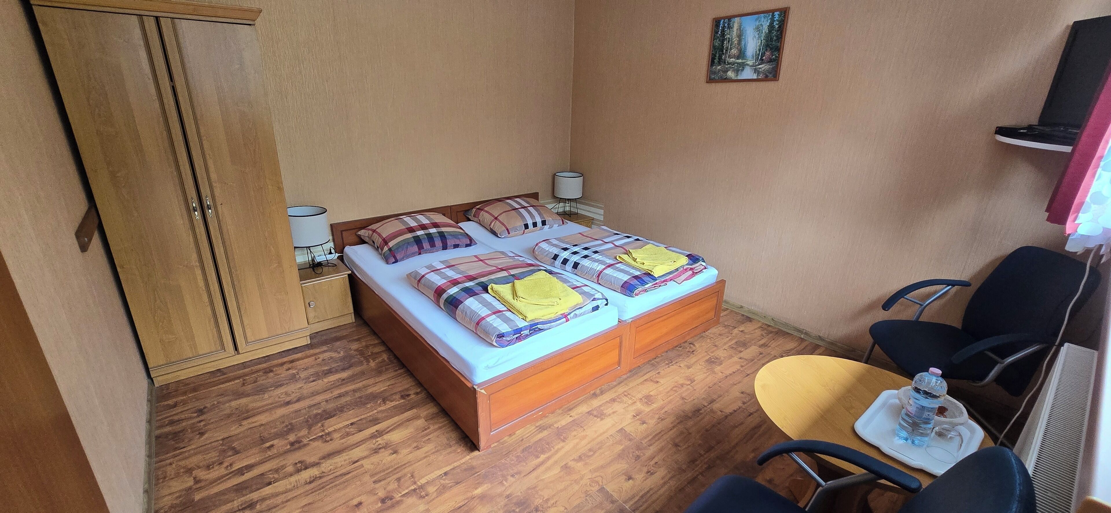 A hotel room with a wooden bed, two bedside tables, a chair, a small table with a water bottle, and a painting on the wall.