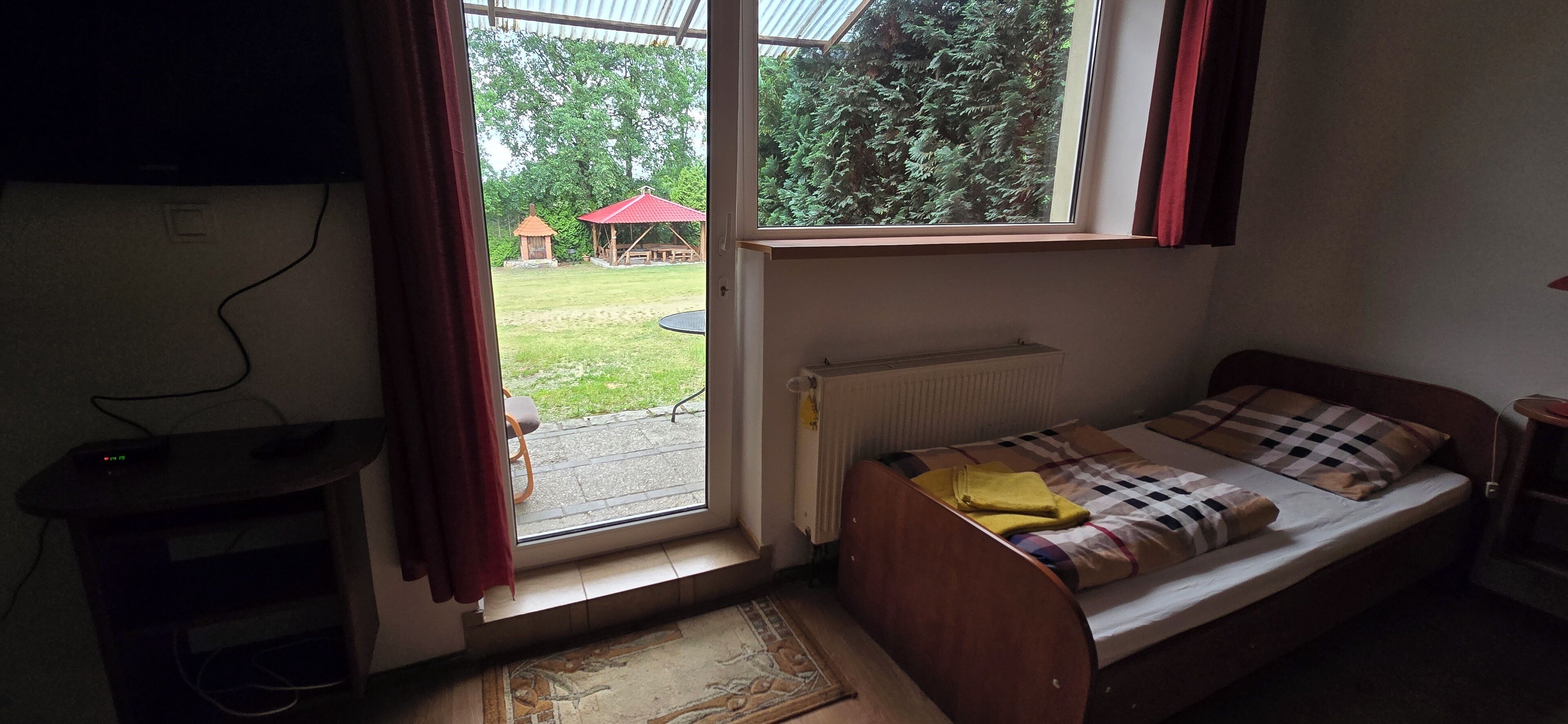 Une petite chambre avec un lit simple, dotée d’une fenêtre donnant sur un jardin. On peut voir un radiateur sous la fenêtre, et un bureau avec une chaise est placé contre le mur.