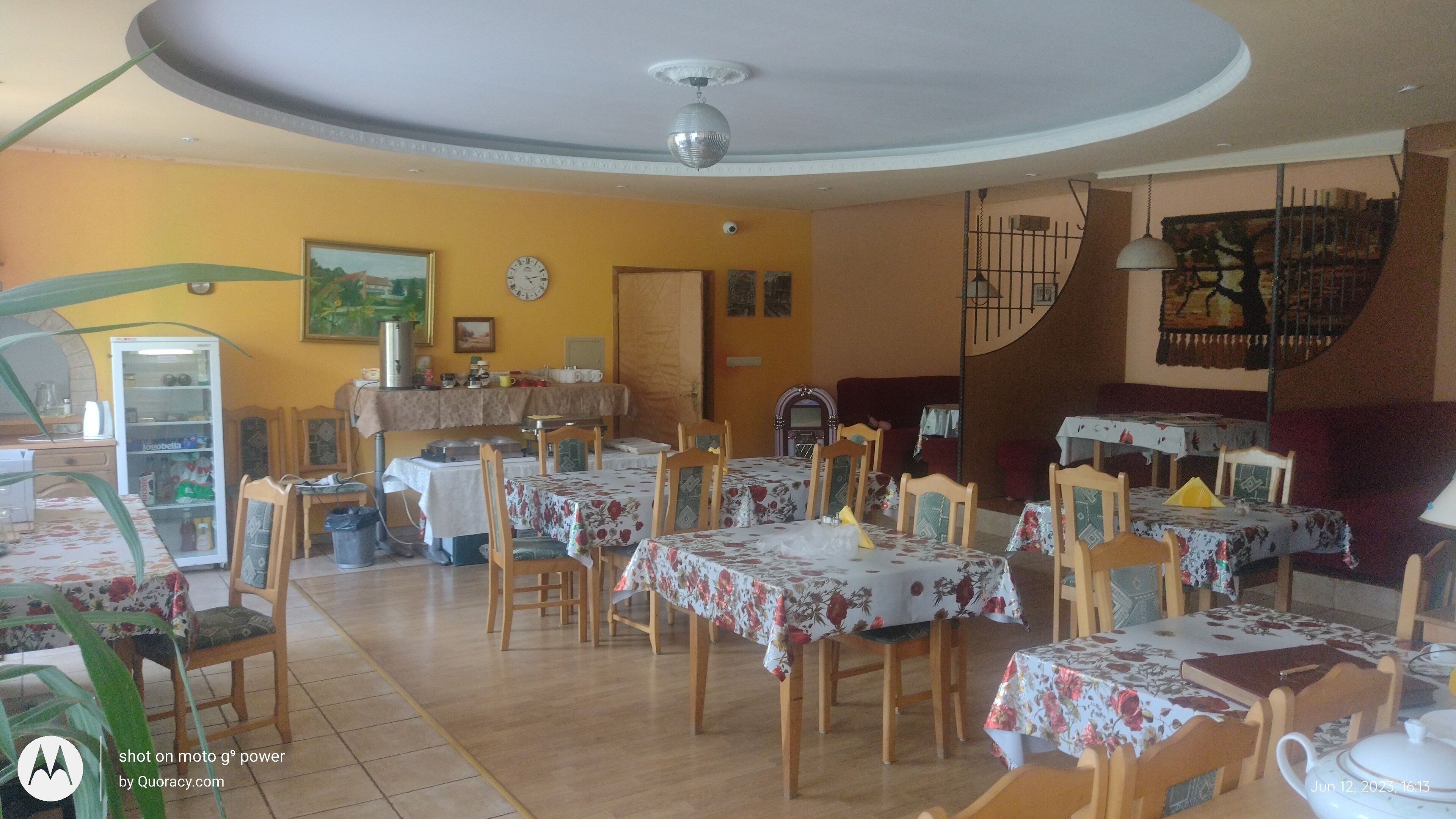 Een eethoek met meerdere tafels en stoelen, een koelkast, een klok en een plafondventilator.