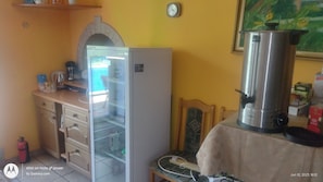 Kitchen area with refrigerator, coffee maker, kettle, and microwave oven, featuring a table and a framed picture.
