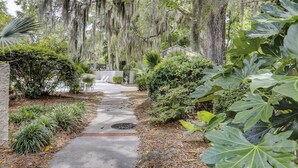 A pathway leading to a pool area surrounded by lush greenery and trees with hanging moss. - Sunset Rentals | Continental Club 16 (Hilton Head Island)