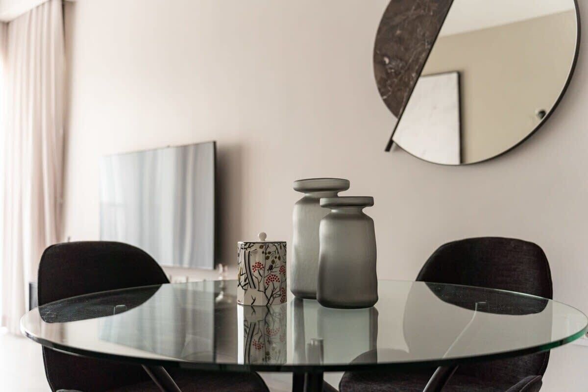 A modern dining area with a glass table, two black chairs, a decorative vase, and a wall-mounted mirror.