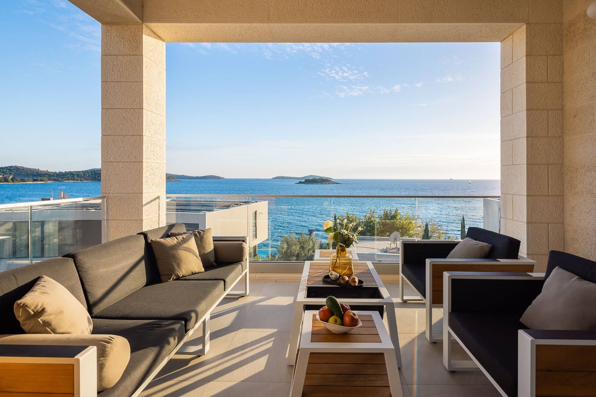 A balcony with a view of the sea, furnished with a sofa, chairs, and a table with fruit.