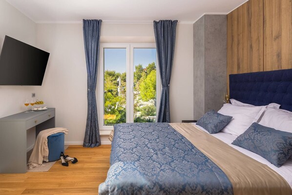A bedroom with a bed, a TV mounted on the wall, a desk with a chair, and a window with curtains.
