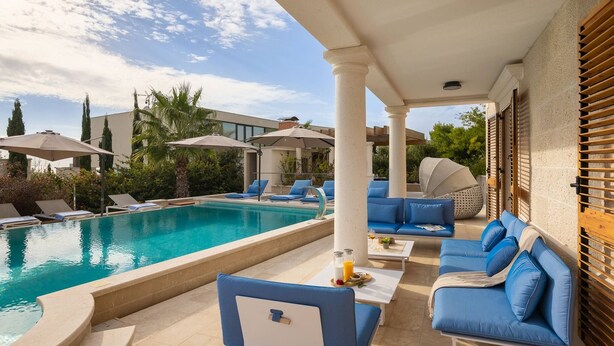 A poolside area with lounge chairs and a table, a clear blue pool, and a covered patio with columns.