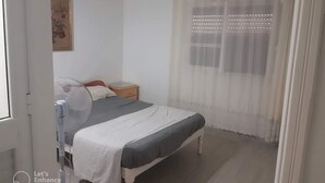 A bedroom with a bed, bedside table, and a window with sheer curtains.
