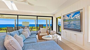 Una sala de estar moderna con un sofá grande, una mesa de centro y un televisor montado en la pared. La habitación tiene ventanas amplias con vistas al océano.