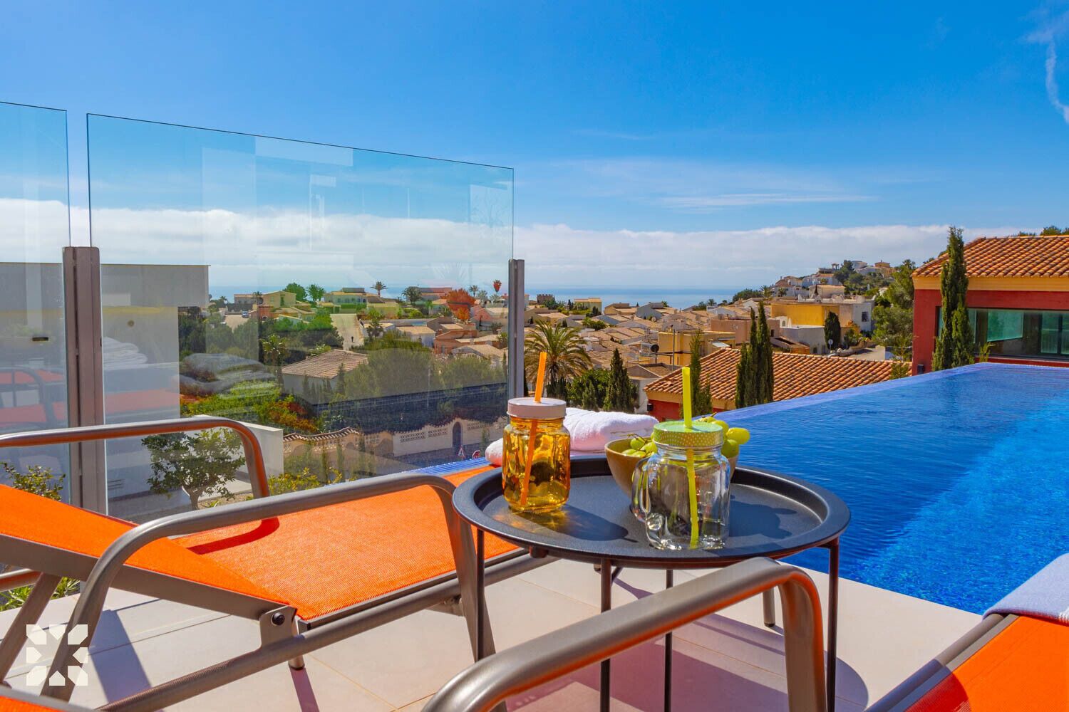 Un espace aménagé au bord de la piscine, doté d’une balustrade en verre, de chaises longues orange et d’une table avec des boissons et des condiments.