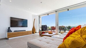 Ein modernes Wohnzimmer mit einem Flachbildfernseher, einer Couch, einem Couchtisch und einem großen Fenster mit Blick ins Freie.