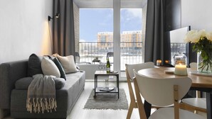 A modern living room with a grey sofa, a small table with a bottle and glasses, and a view of buildings through large windows. - Elite Apartments Stępkarska Premium (Gdansk)