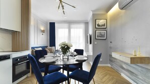 A modern kitchen with a built-in oven, a dining area with a round table and blue chairs, and a living area with a TV and artwork on the wall. - Elite Apartments Stępkarska Premium (Gdansk)