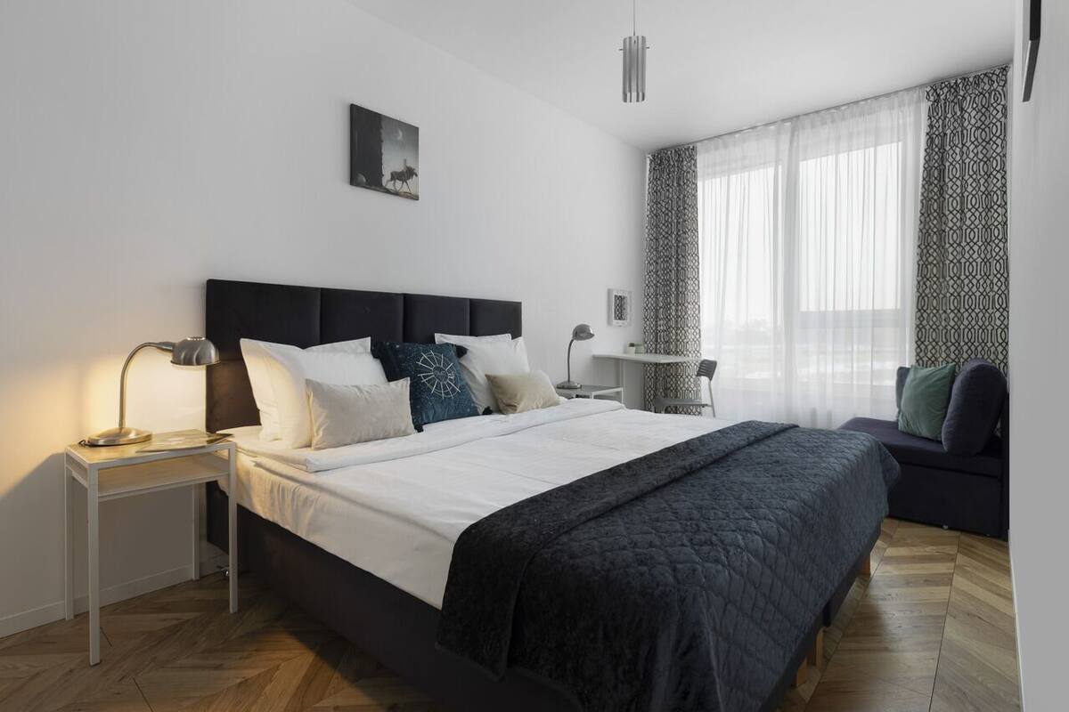 Un dormitorio moderno con una cama grande, mesitas de noche, un pequeño sofá y una ventana con cortinas translúcidas.
