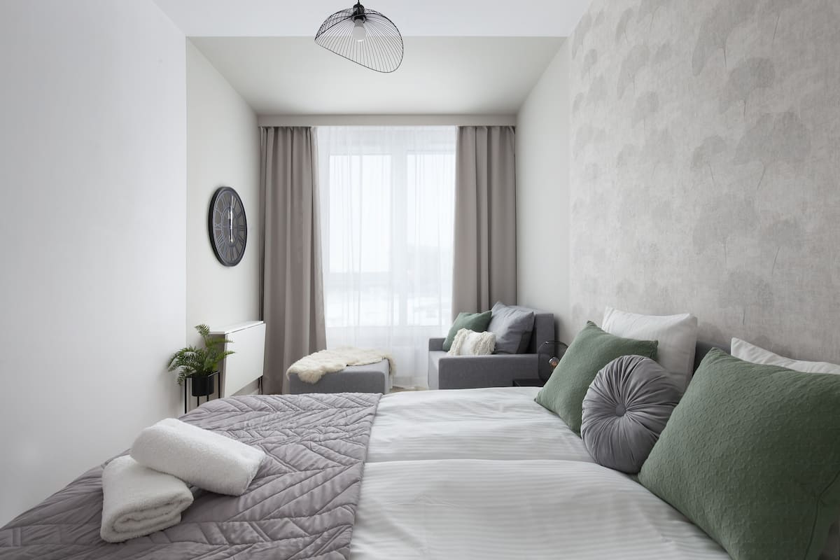 Un dormitorio moderno con una cama grande, una mesita de noche, una silla, una ventana con cortinas y un reloj en la pared.
