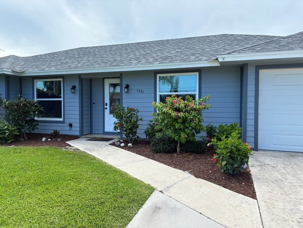 A single-story house with a grey roof, a front door, and a garage. There are small shrubs and a larger bush with pink flowers in front of the house. - Golf, Tennis, Bike & Beach in Hobe Sound (Hobe Sound)