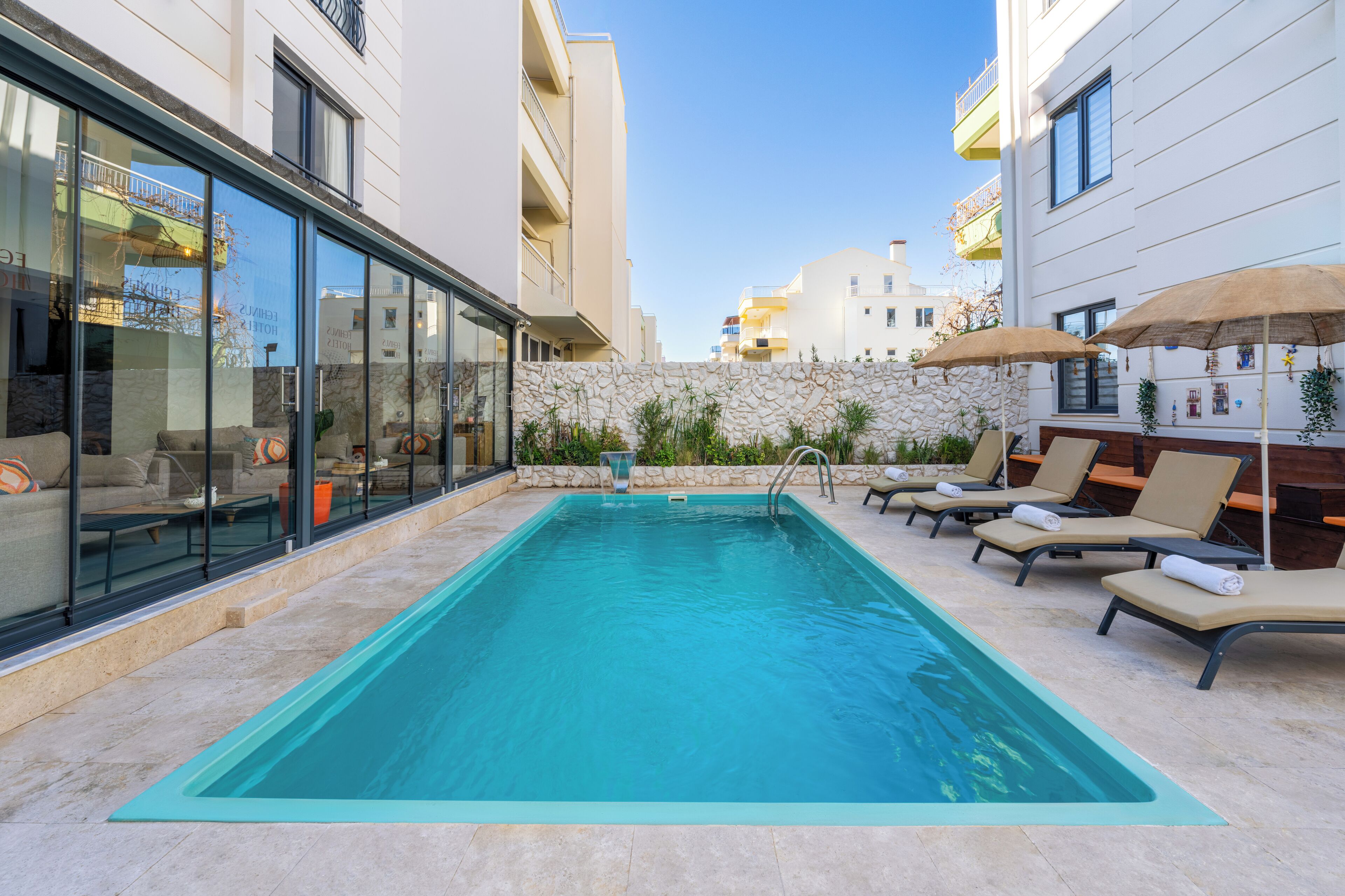 A swimming pool with lounge chairs and umbrellas, surrounded by stone walls and modern buildings.