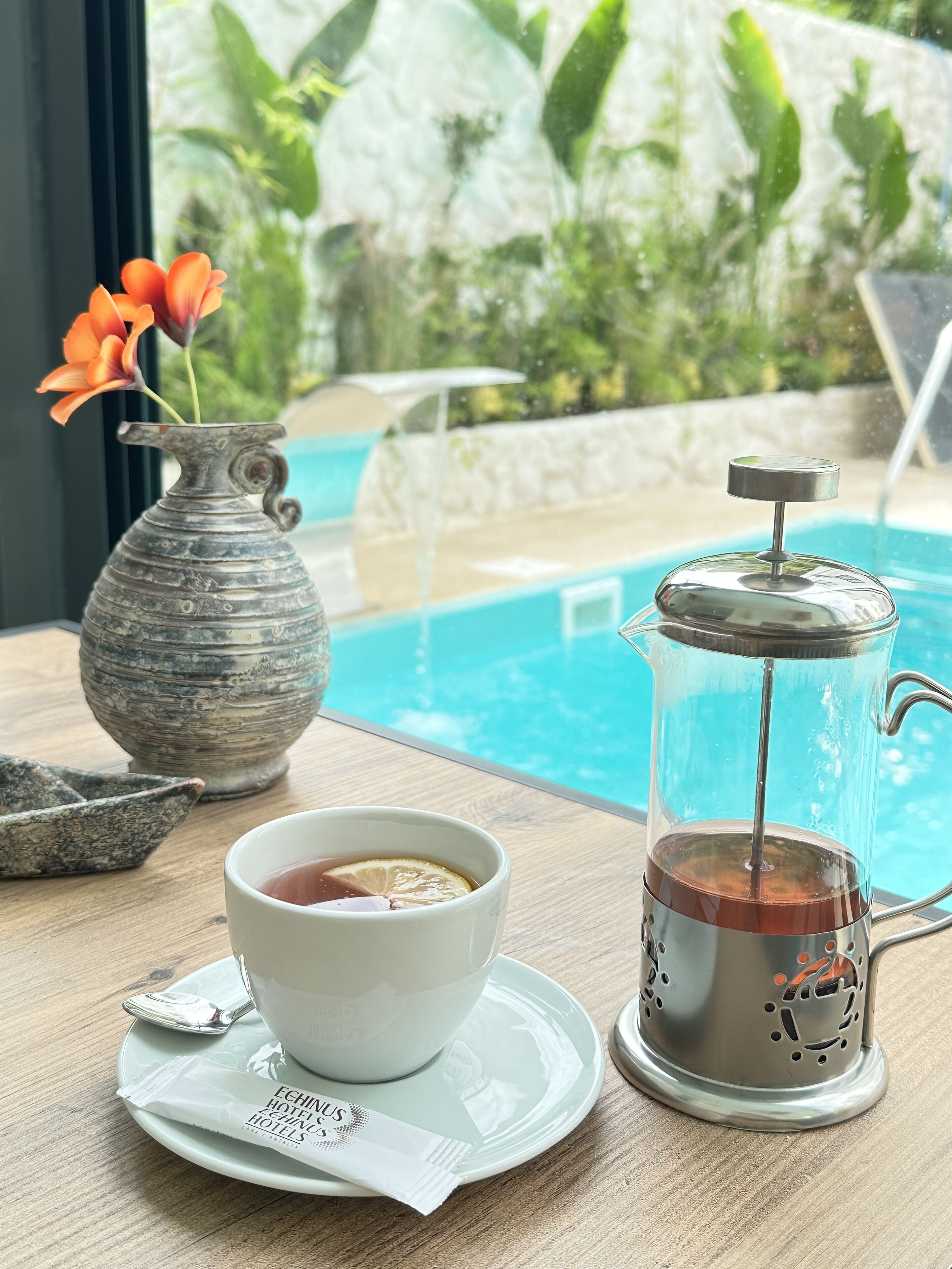 A table with a coffee pot, a cup of tea, a flower vase, and a view of a pool.