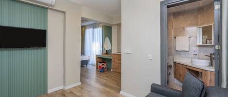 A modern hotel room with a flat-screen TV, wooden flooring, a grey sofa, and a bathroom with a large mirror and sink.