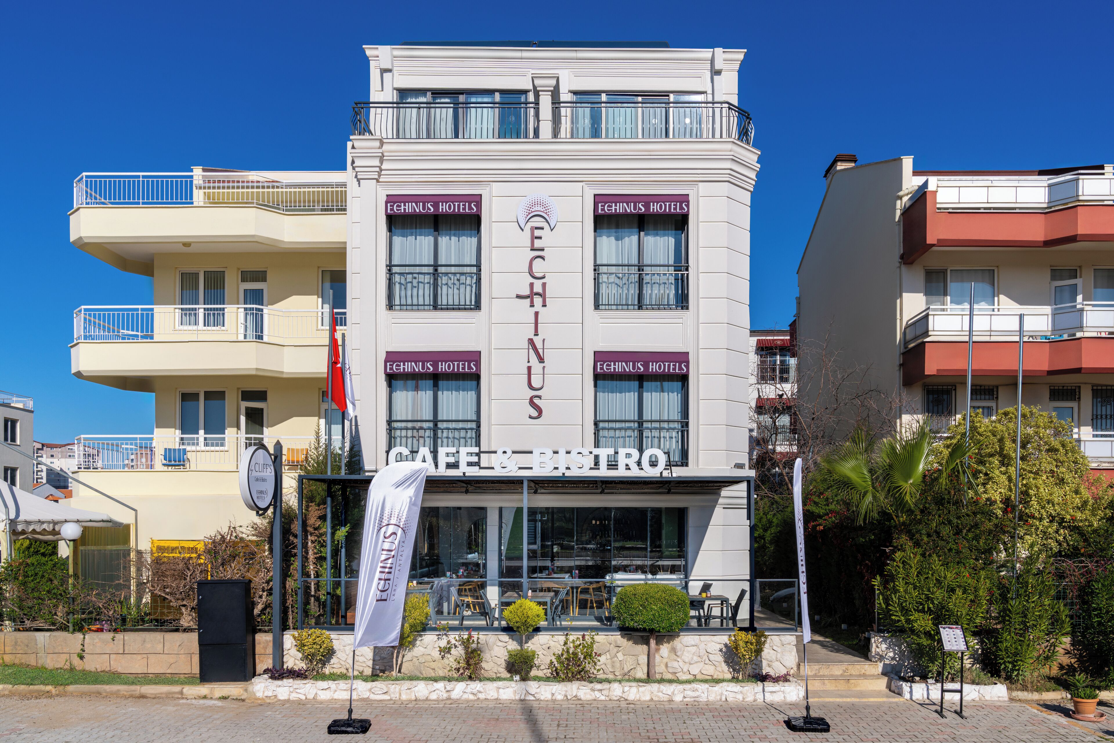 Un edificio con la señalización 'ECHINUS HOTELS' y 'CAFÉ & BISTRO'. Cuenta con balcones, un área de café con mesas al aire libre y fachada de piedra.