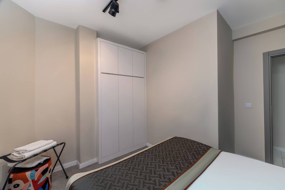 A bedroom with a bed, a wardrobe, a small ironing board, and a colorful bag.