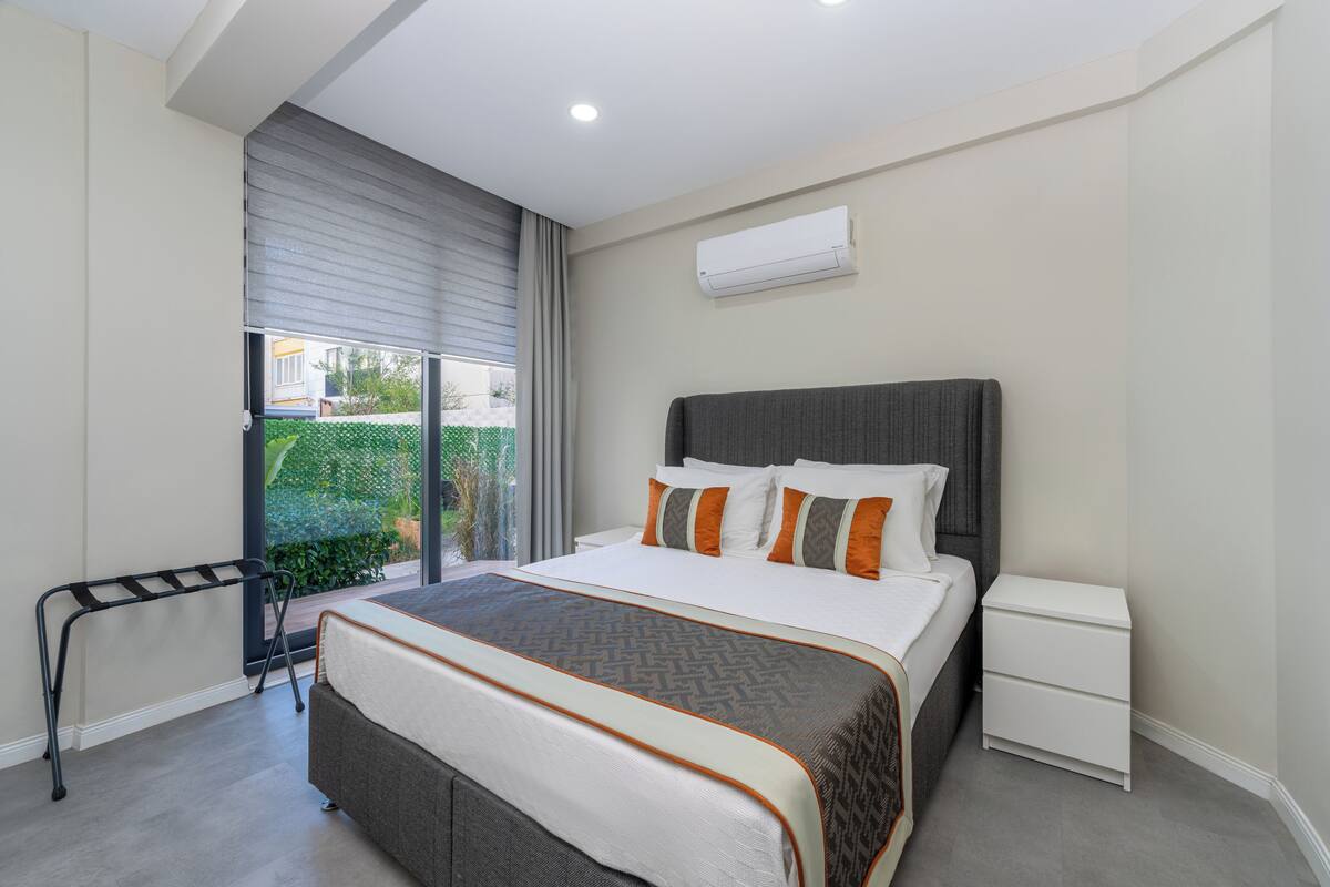 A modern bedroom with a large bed, a nightstand, a window with a view, and an air conditioning unit.