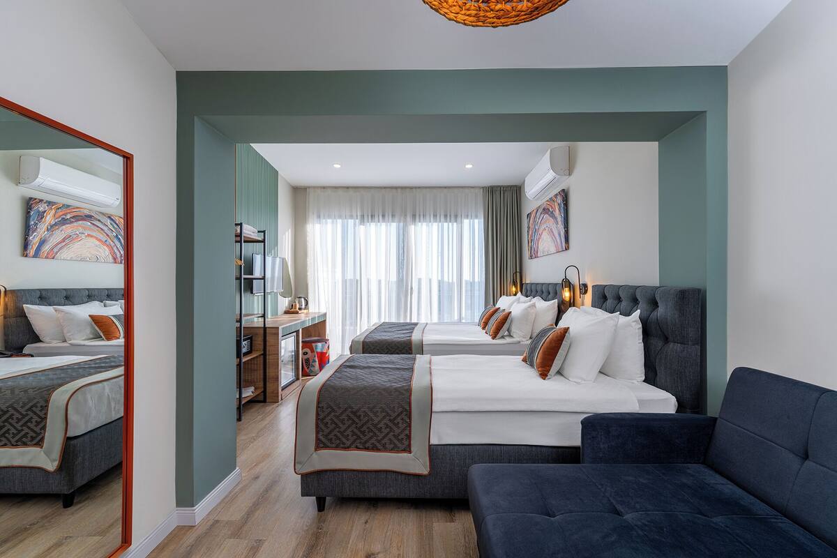 A modern hotel room with two beds, a sofa, a desk, and a large mirror.