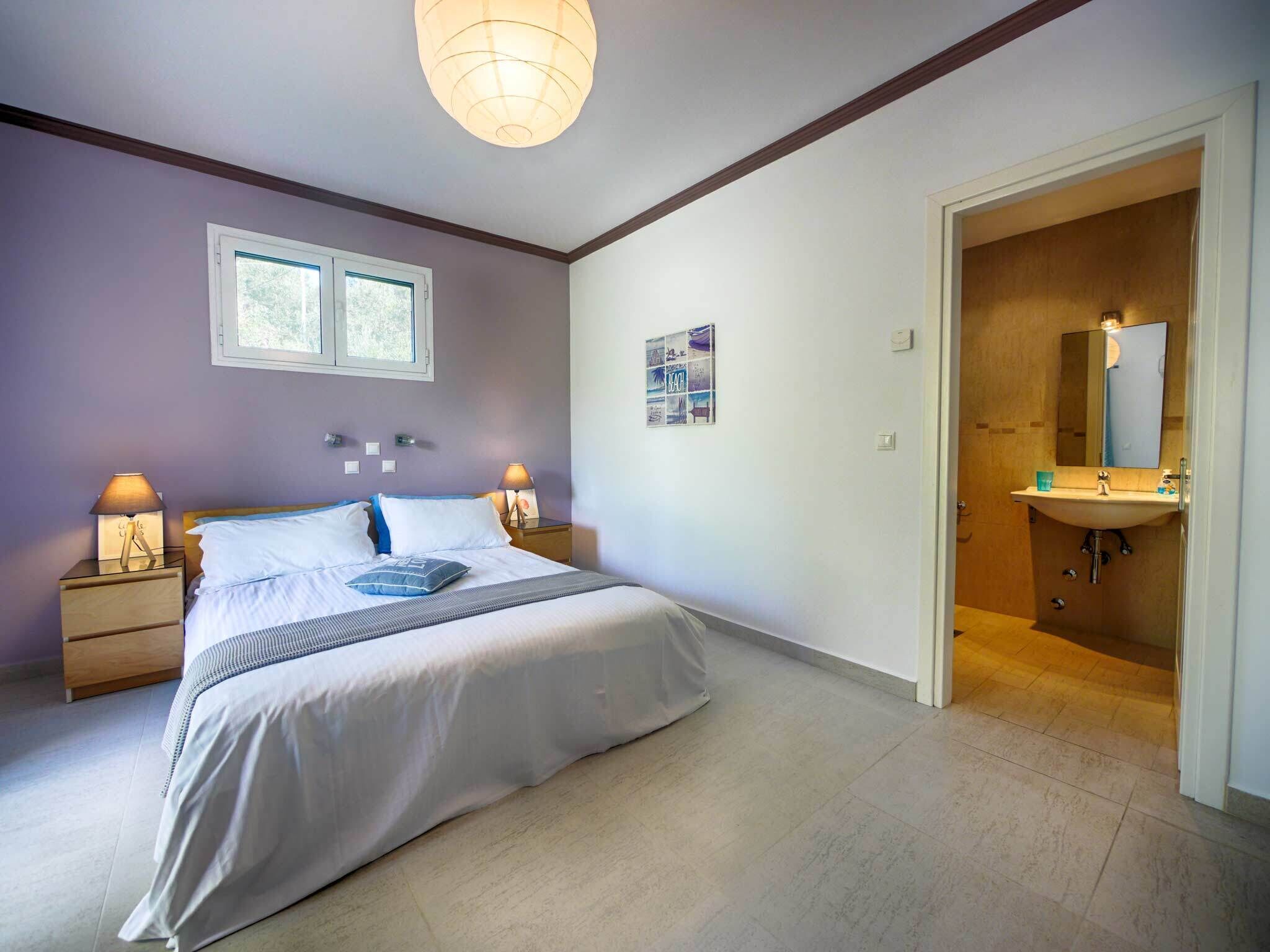A bedroom with a bed, bedside lamps, a window, and a bathroom with a sink and mirror.