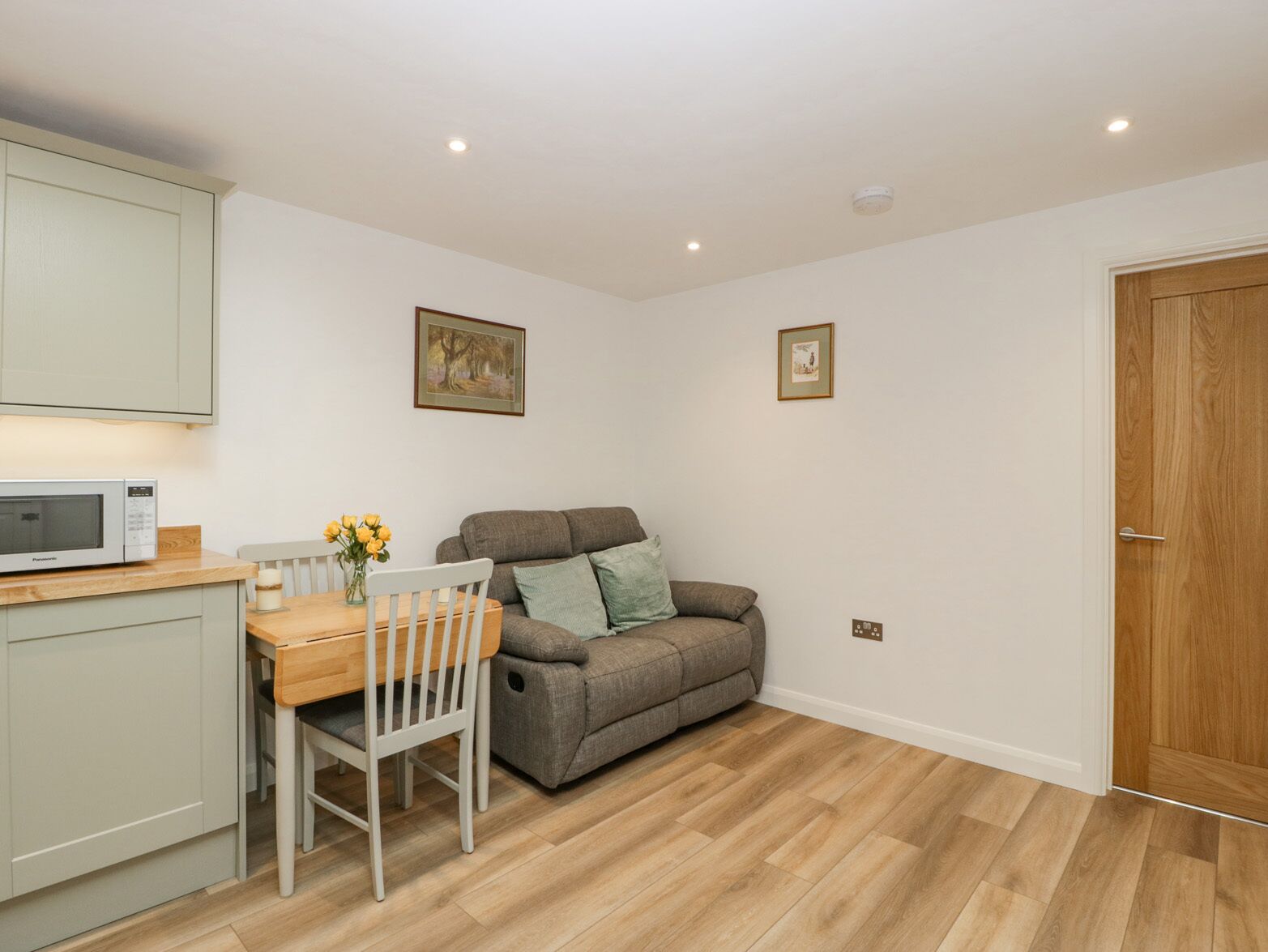 A compact living space with a kitchenette, a dining table with chairs, a grey sofa, and a wooden door.
